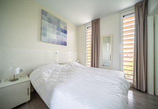 Helles Schlafzimmer im Pavilion Waterfront, EuroParcs Veluwemeer, mit Doppelbett, Kunstwerk und Fenstern.