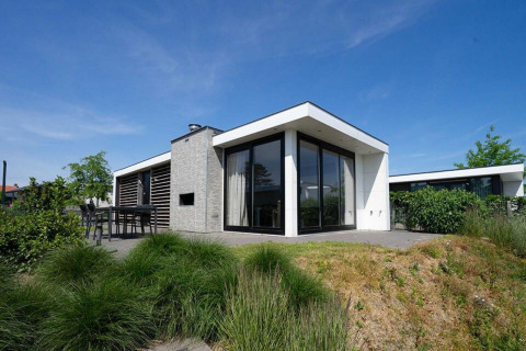 Modernes Pavilion Waterfront-Lodge mit großen Fenstern, Terrasse und Garten unter blauem Himmel.
