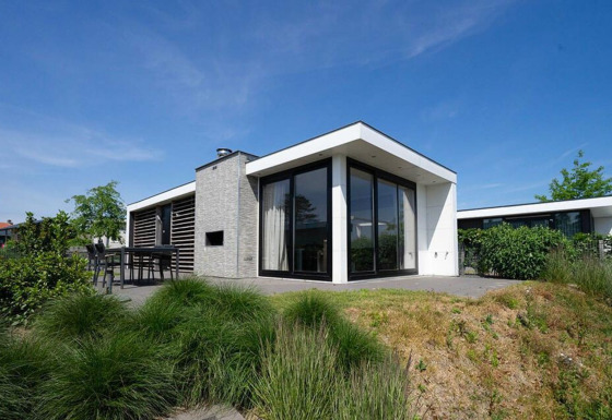 Modern Pavilion Waterfront lodge featuring large glass windows, patio, and lush garden under blue sky.