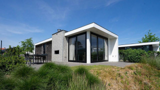Modernes Pavilion Waterfront-Lodge mit großen Fenstern, Terrasse und Garten unter blauem Himmel.