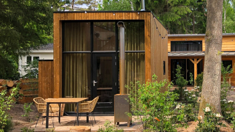 Casa pequeña moderna de madera con ventana grande, terraza con sillas y mesa en entorno boscoso.