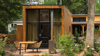 Casa pequeña moderna de madera con ventana grande, terraza con sillas y mesa en entorno boscoso.