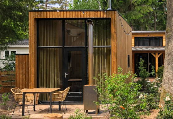 Casa pequeña moderna de madera con ventana grande, terraza con sillas y mesa en entorno boscoso.