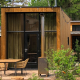 Modern wooden tiny house with large glass windows, patio seating, and garden in a lush forest setting.