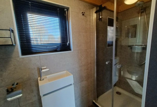Baño moderno con ducha de vidrio, lavabo pequeño y ventana con persianas en Tiny House de EuroParcs Veluwemeer.