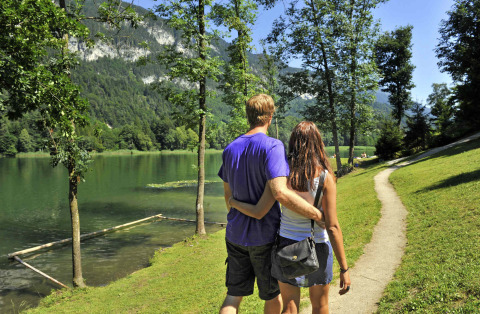 Pareja caminando junto a un lago rodeado de montañas y árboles en Camping Seeblick Toni, Tirol, Austria.