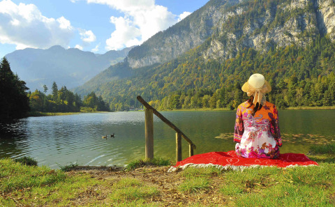 Mujer con sombrero sentada en manta junto al lago y montañas en Camping Seeblick Toni, Tirol, Austria.