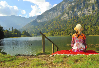 Kvinde med hat sidder på tæppe ved sø i bjergene, Camping Seeblick Toni, Tirol, Østrig, solrig dag.
