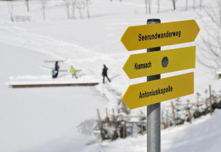 Winterlandschaft in Tirol mit gelbem Wegweiser bei Camping Seeblick Toni und Wanderern im Schnee.