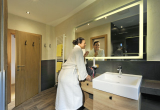 Woman in bathrobe brushing teeth at the sink in a modern bathroom at Camping Seeblick Toni in Tirol, Austria.