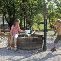 Twee kinderen spelen met een waterpomp en houten ton op Camping Seeblick Toni in Tirol, Oostenrijk.