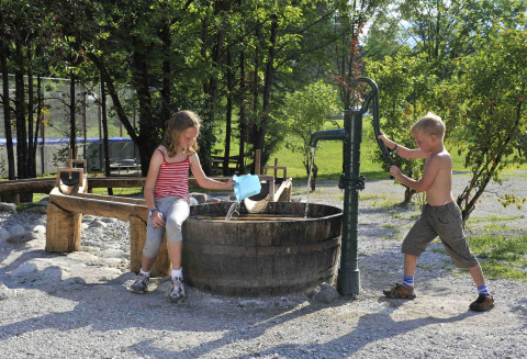 Dos niños juegan con una bomba de agua antigua junto a un barril en Camping Seeblick Toni, Tirol, Austria.