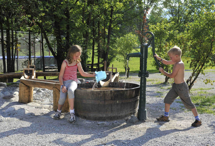 Zwei Kinder spielen an einer alten Wasserpumpe bei einer Holzbottich auf Camping Seeblick Toni in Tirol.