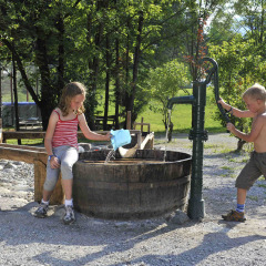 Twee kinderen spelen met een waterpomp en houten ton op Camping Seeblick Toni in Tirol, Oostenrijk.