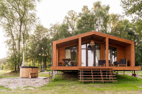 Træhytte kaldet Lakehouse med hottub foran og terrasse, omgivet af grønne træer og natur.