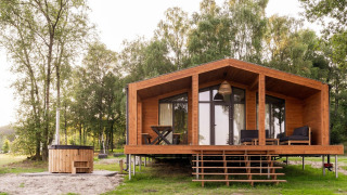 Træhytte kaldet Lakehouse med hottub foran og terrasse, omgivet af grønne træer og natur.