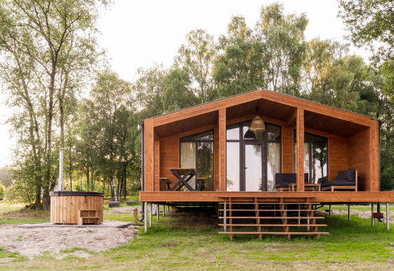 Træhytte kaldet Lakehouse med hottub foran og terrasse, omgivet af grønne træer og natur.