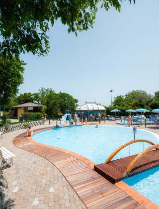 Outdoor swimming pool with wooden bridge and sun loungers at Camping Centro Vacanze San Marino, Italy, on a sunny day.