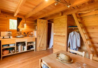 Interno di una rustica Cahutte glamping con angolo cucina, tavolo da pranzo e scala per il soppalco.