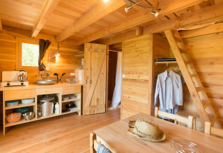 Interior de una acogedora Cahutte glamping con cocina, comedor, armario abierto y escalera al altillo.