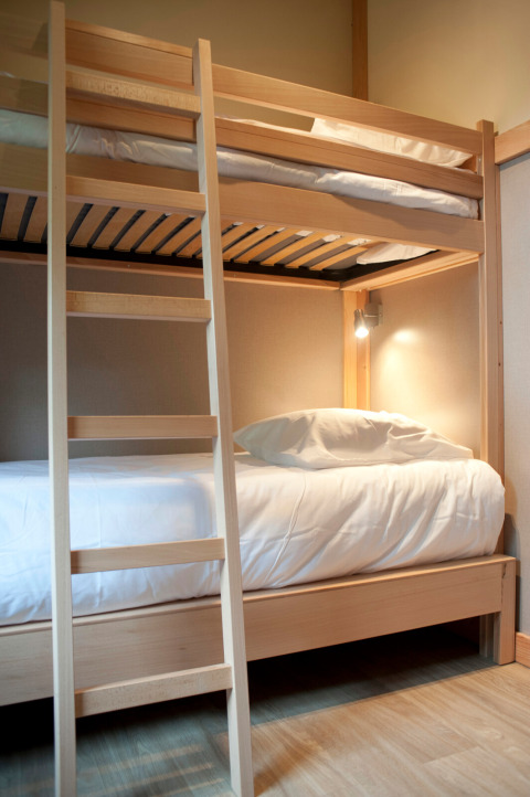 Stapelbed van licht hout met wit beddengoed in een glamping Chalet Montana kamer, lamp aan de muur.