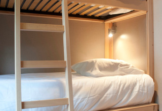 Helle Holzetagenbetten mit weißer Bettwäsche im Glamping Chalet Montana, gemütliche Beleuchtung an der Wand.