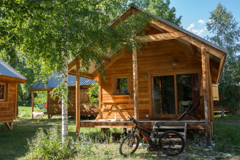 Cabaña de madera y bicicleta frente al Chalet Montana en Huttopia Vallouise, Francia, rodeados de árboles verdes.