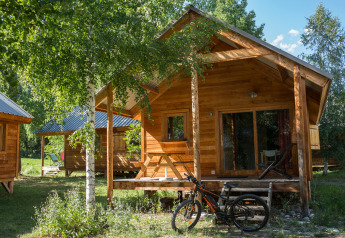 Træchalet og cykel foran Chalet Montana i Huttopia Vallouise, Frankrig, omgivet af sommergrønne træer.