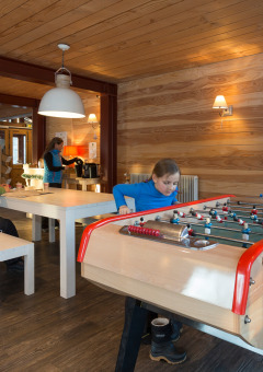 Niños juegan al futbolín mientras adultos descansan en la sala común de Les chalets Huttopia de Bourg-St-Maurice, Francia.