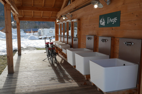 Udendørs vaskestation med flere vaske og to cykler på en træterrasse, sne og campingvogne i baggrunden.