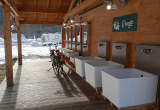 Udendørs vaskestation med flere vaske og to cykler på en træterrasse, sne og campingvogne i baggrunden.