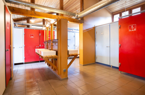 Baño comunitario moderno con puertas rojas y blancas, lavabos y luz natural en Huttopia Bourg-St-Maurice.