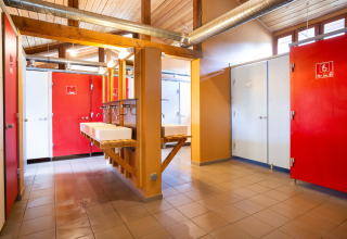 Baño comunitario moderno con puertas rojas y blancas, lavabos y luz natural en Huttopia Bourg-St-Maurice.