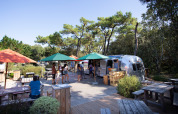 Udendørs caféområde med foodtruck, parasoller og borde blandt træer i Huttopia Noirmoutier, Pays de la Loire.