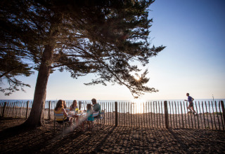 Gezin dineert onder een boom bij Huttopia Noirmoutier, terwijl een loper langs het strand passeert.
