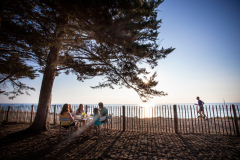 Familia disfruta de una comida bajo un árbol en Huttopia Noirmoutier, mientras una persona corre en la playa.