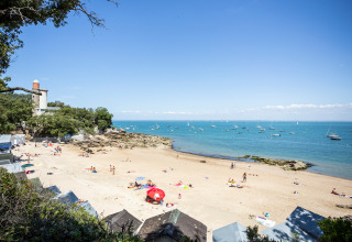 Escena de playa en Huttopia Noirmoutier, Pays de la Loire, Francia, con personas y barcos en el agua.