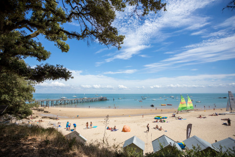 Strandscene ved Huttopia Noirmoutier med sejlbåde og besøgende på sandet under en klar blå himmel.