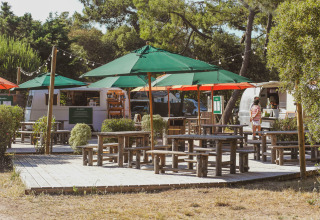 Outdoor seating area with green umbrellas and wooden benches by food trucks at Huttopia Noirmoutier, France.