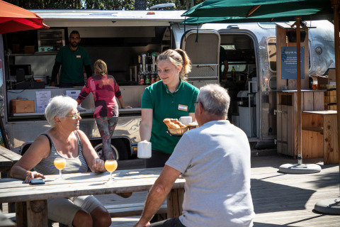 Personal sirve desayuno a dos huéspedes sentados al aire libre frente a un food truck en Huttopia Noirmoutier.