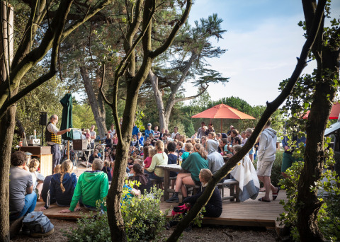 Een grote groep bekijkt een buitenoptreden onder de bomen bij Huttopia Noirmoutier vakantiepark in Frankrijk.
