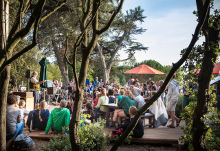 Een grote groep bekijkt een buitenoptreden onder de bomen bij Huttopia Noirmoutier vakantiepark in Frankrijk.