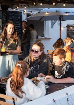 Una familia disfruta de una comida al aire libre mientras una camarera sirve bebidas en Huttopia Ars-en-Ré, Francia.