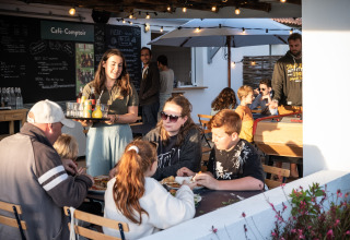 En glad familie nyder et måltid udendørs, mens en servitrice serverer drikkevarer på Huttopia Ars-en-Ré, Frankrig.