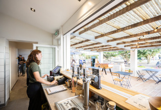 Kvinde bag disken i en café ved Huttopia Ars-en-Ré med udsigt til udendørs siddepladser og bordfodbold.