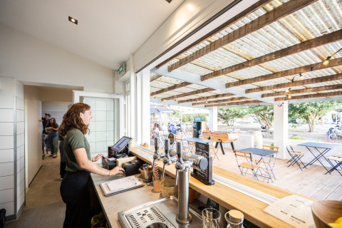 Woman works behind the counter at Huttopia Ars-en-Ré café overlooking outdoor seating and foosball table.