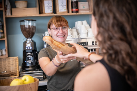 Una empleada sonriente en Huttopia Ars-en-Ré entrega una baguette a una huésped en el parque vacacional.