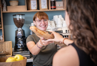 En smilende medarbejder i Huttopia Ars-en-Ré overleverer en baguette til en gæst i ferieparken.