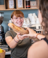 Una empleada sonriente en Huttopia Ars-en-Ré entrega una baguette a una huésped en el parque vacacional.