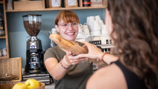 Una empleada sonriente en Huttopia Ars-en-Ré entrega una baguette a una huésped en el parque vacacional.